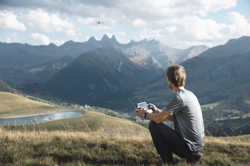 Télépilote pilotant son drone au col de la Chal, la retenue collinaire en contrebas et les Aiguilles d'Arves à l'horizon